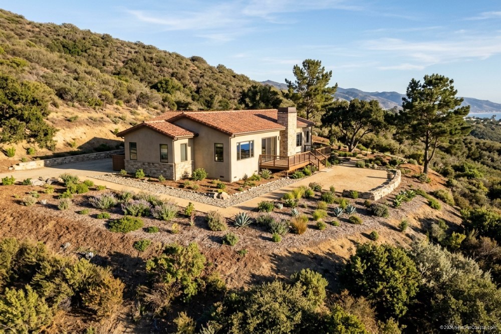 Photorealistic DSLR photograph of a hillside single-family home in coastal California with cleared defensible space, low native plants, gravel pathways, no dense brush, afternoon sunlight, showing proper vegetation management