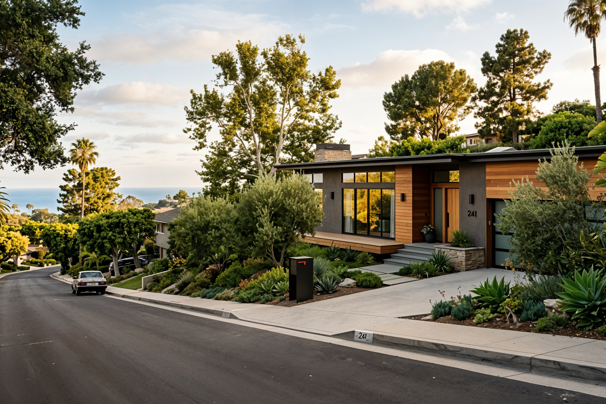 Modern coastal California hillside home with partial ocean view in the distance, tree-lined residential street